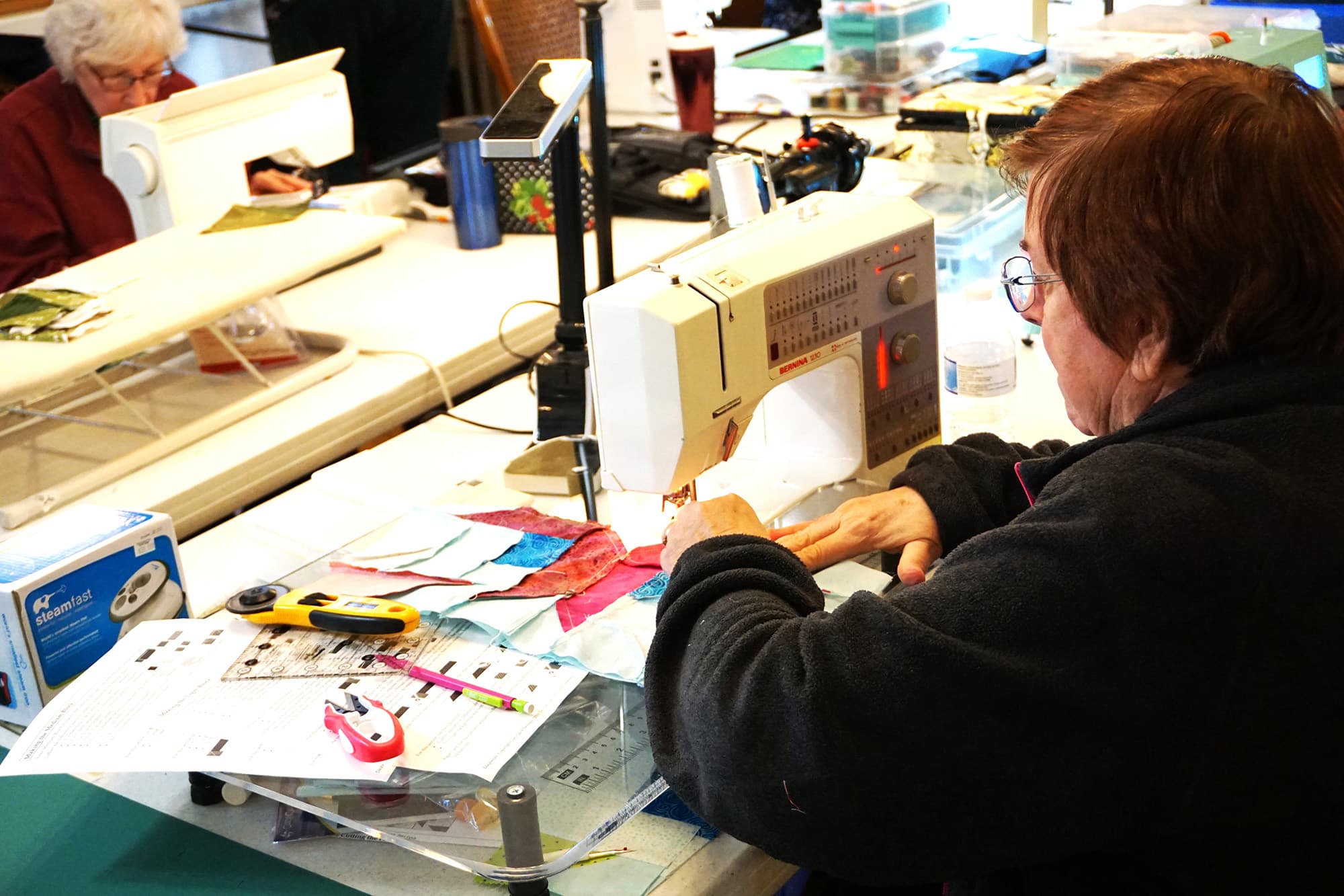 A woman works at a sewing machine surrounded by colorful fabric pieces and sewing tools.