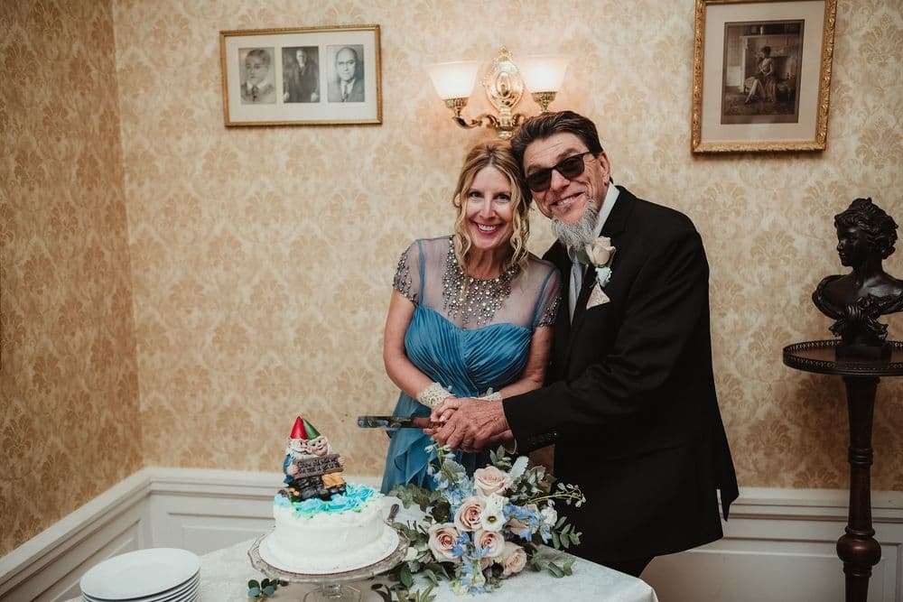 A man and woman, both elegantly dressed, smile as they prepare to cut a wedding cake adorned with flowers and a small figure.