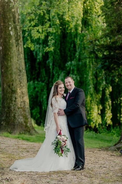 A happy couple in wedding attire poses together outdoors with greenery in the background.