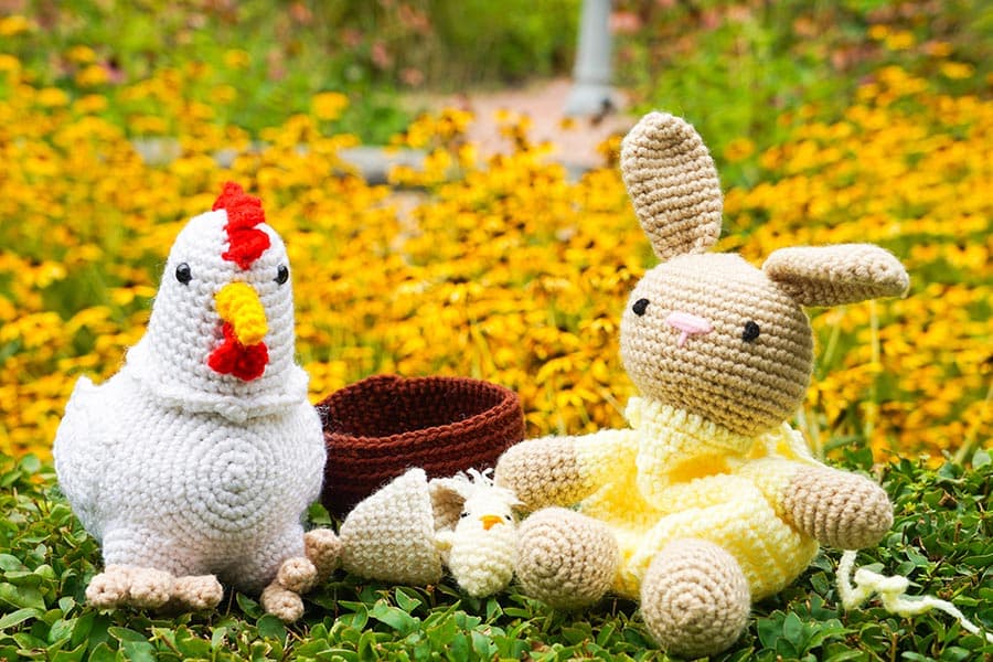 A crocheted chicken and bunny sit together on a grassy patch surrounded by yellow flowers.