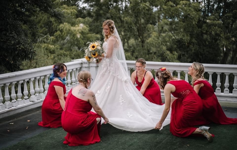 A smiling bride in a white wedding gown poses with her bridal party, dressed in red, as they help adjust her train outdoors.