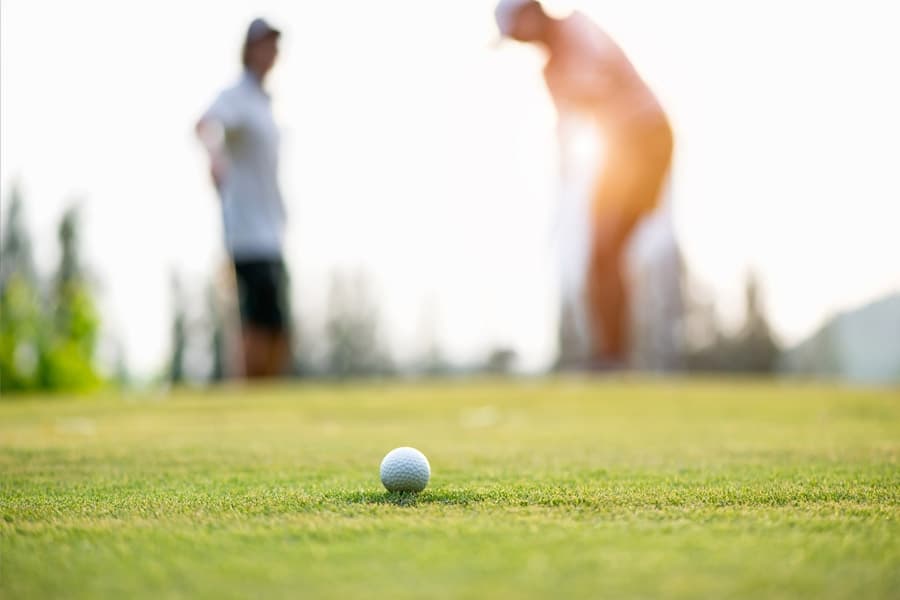 A golf ball sits on the green with two players blurred in the background.