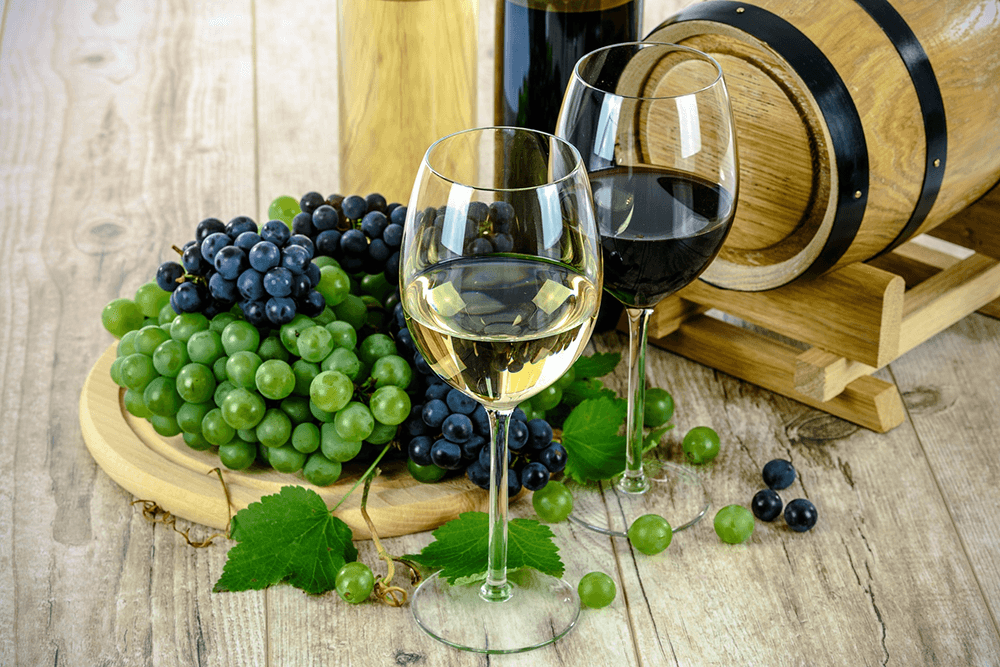 Two wine glasses filled with white and red wine sit beside a platter of green and black grapes, with a wooden barrel in the background.