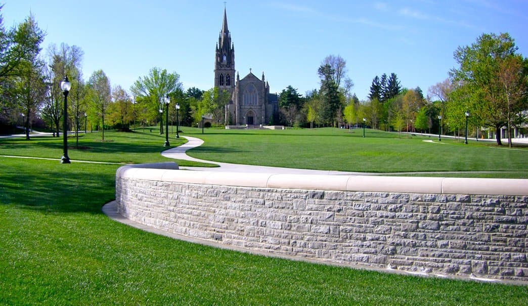 A well-maintained park with a stone wall and a church tower in the background against a clear blue sky.