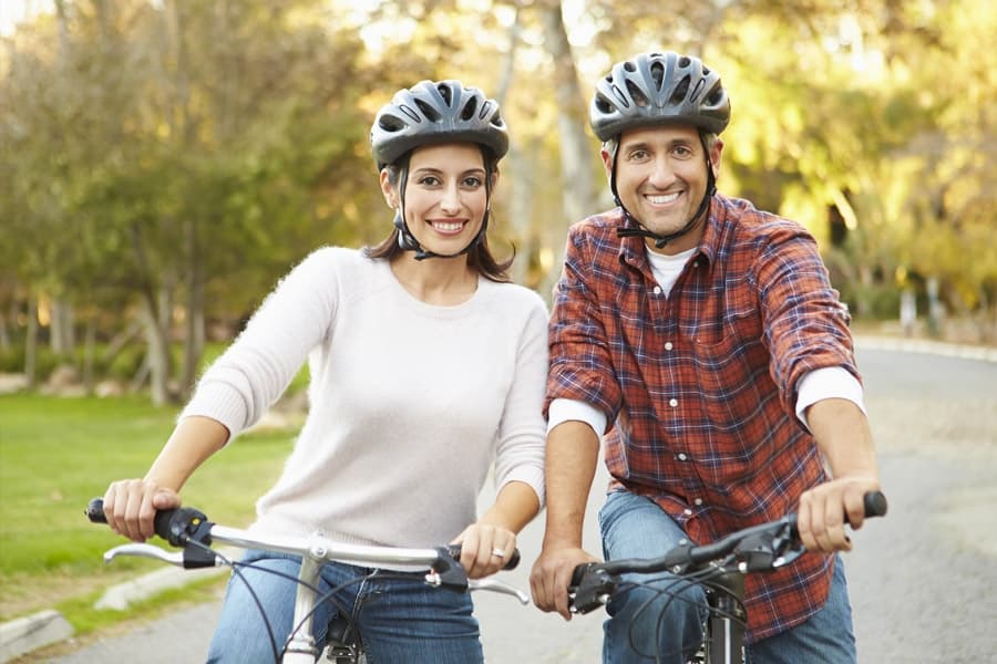 Two smiling people wearing helmets pose on bicycles in a park.