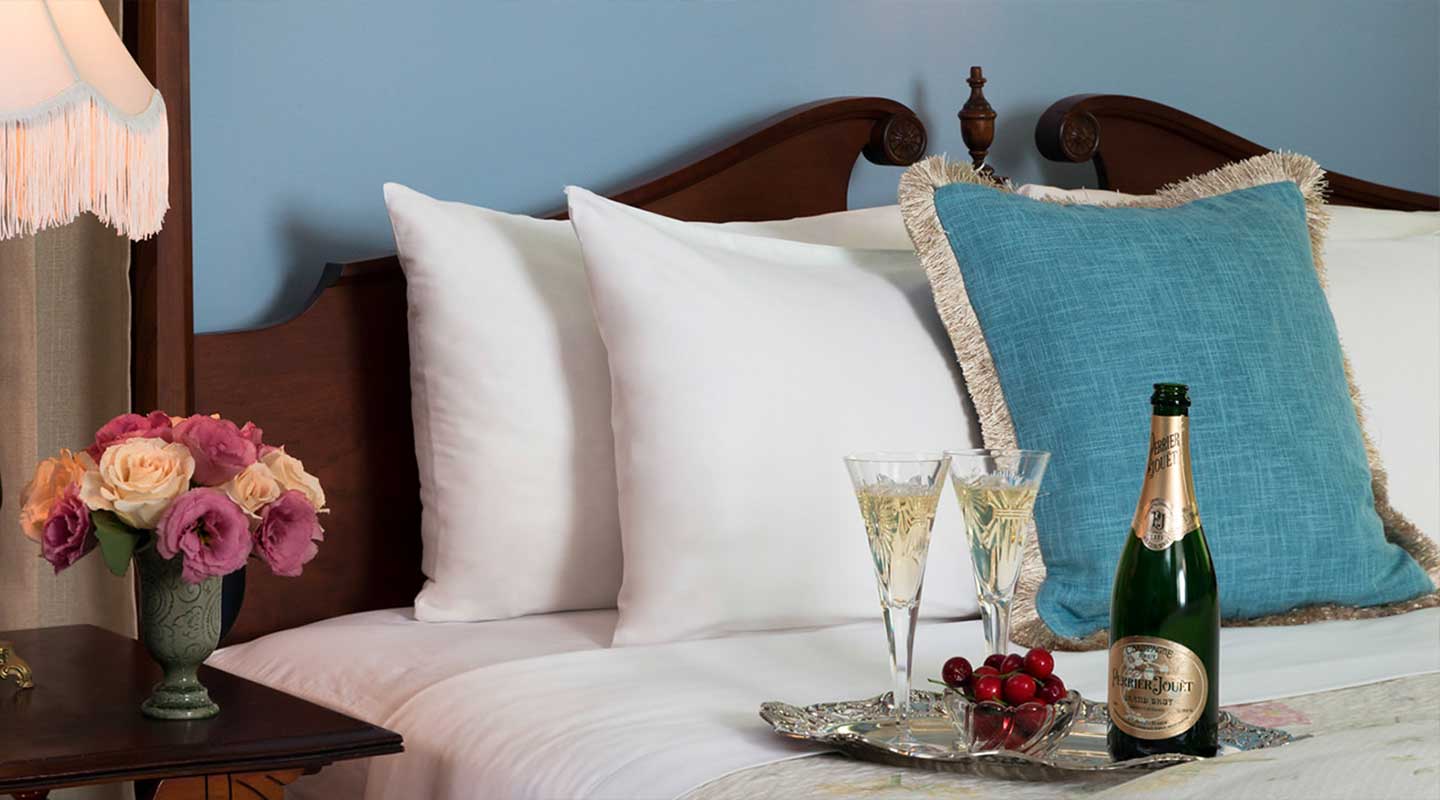 A cozy bed with white pillows, a turquoise accent pillow, a vase of flowers, two champagne glasses, and a bottle of champagne.