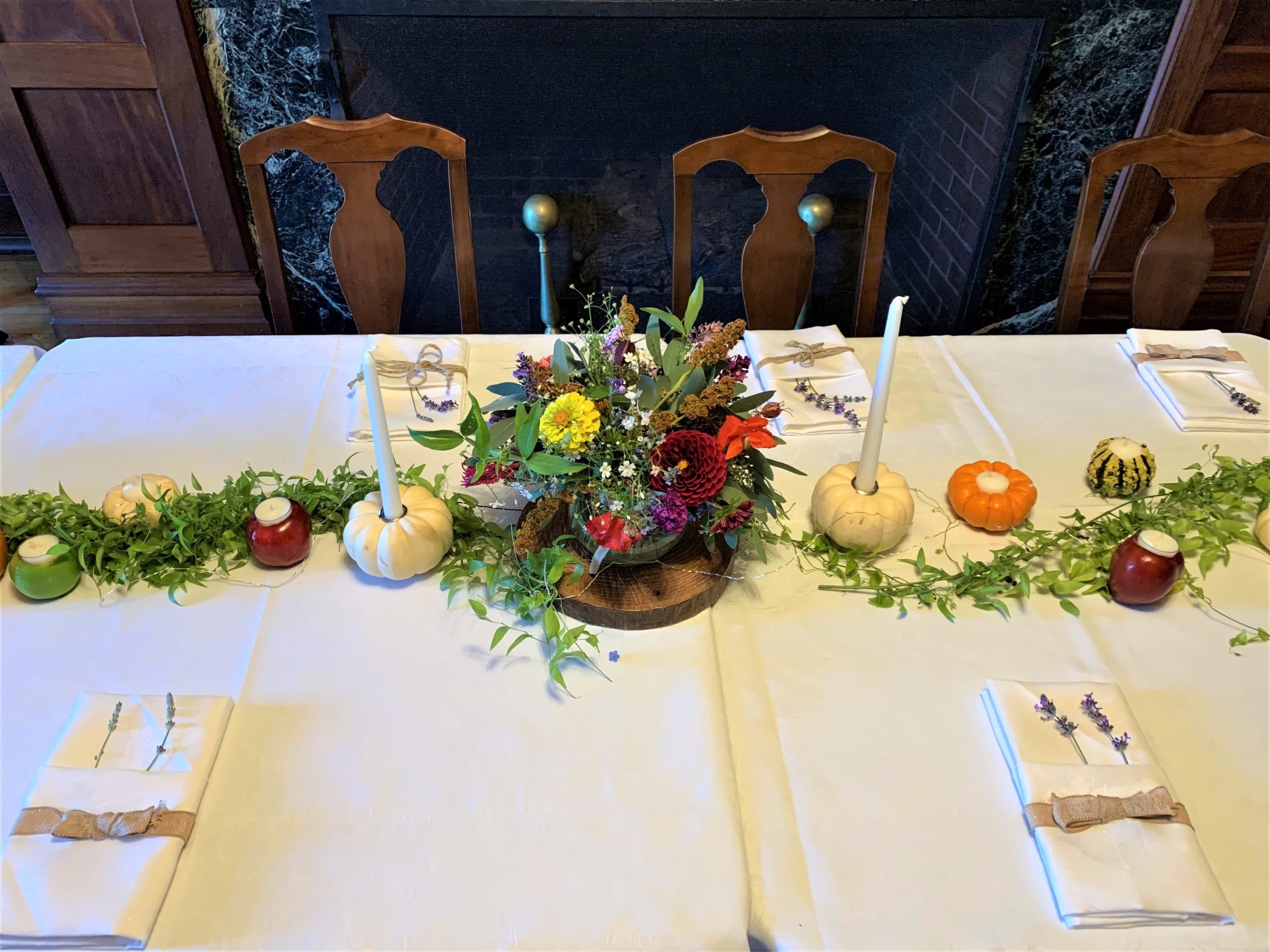 A beautifully set dining table decorated with a floral arrangement, candles, pumpkins, and greenery.