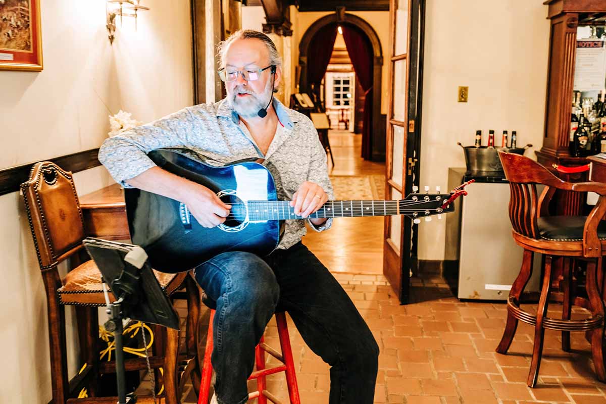 A man with glasses plays an acoustic guitar while seated on a stool in a rustic setting.