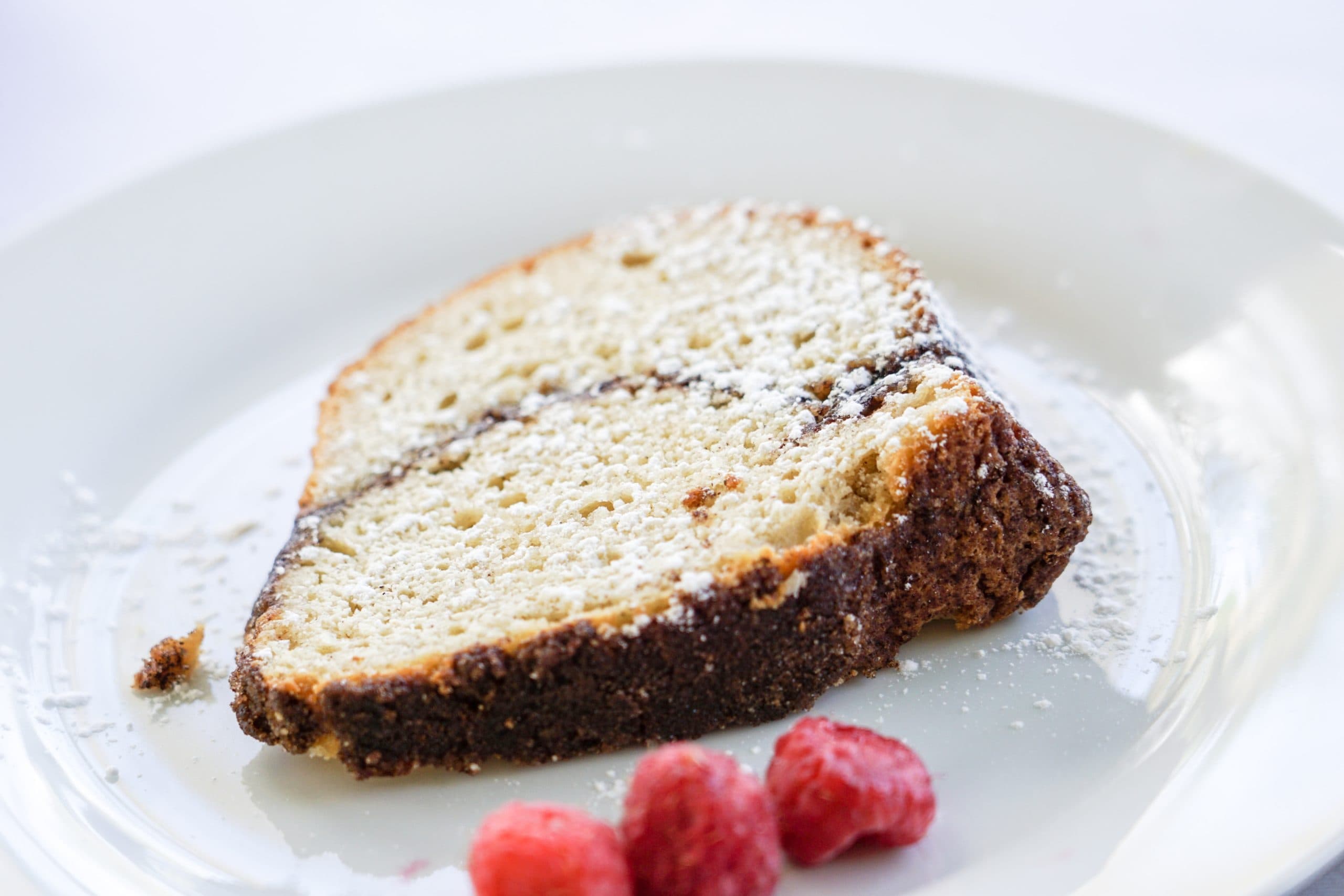 A slice of cake dusted with powdered sugar, accompanied by fresh raspberries on a white plate.