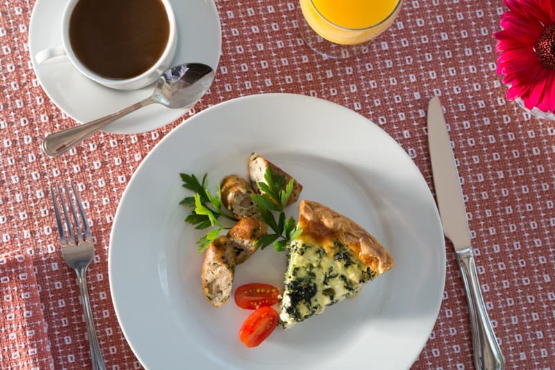A plate with spinach quiche, sausage, cherry tomatoes, a coffee cup, and orange juice, set on a textured tablecloth.