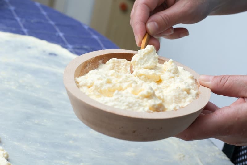 A person stirring fresh cheese in a wooden bowl.