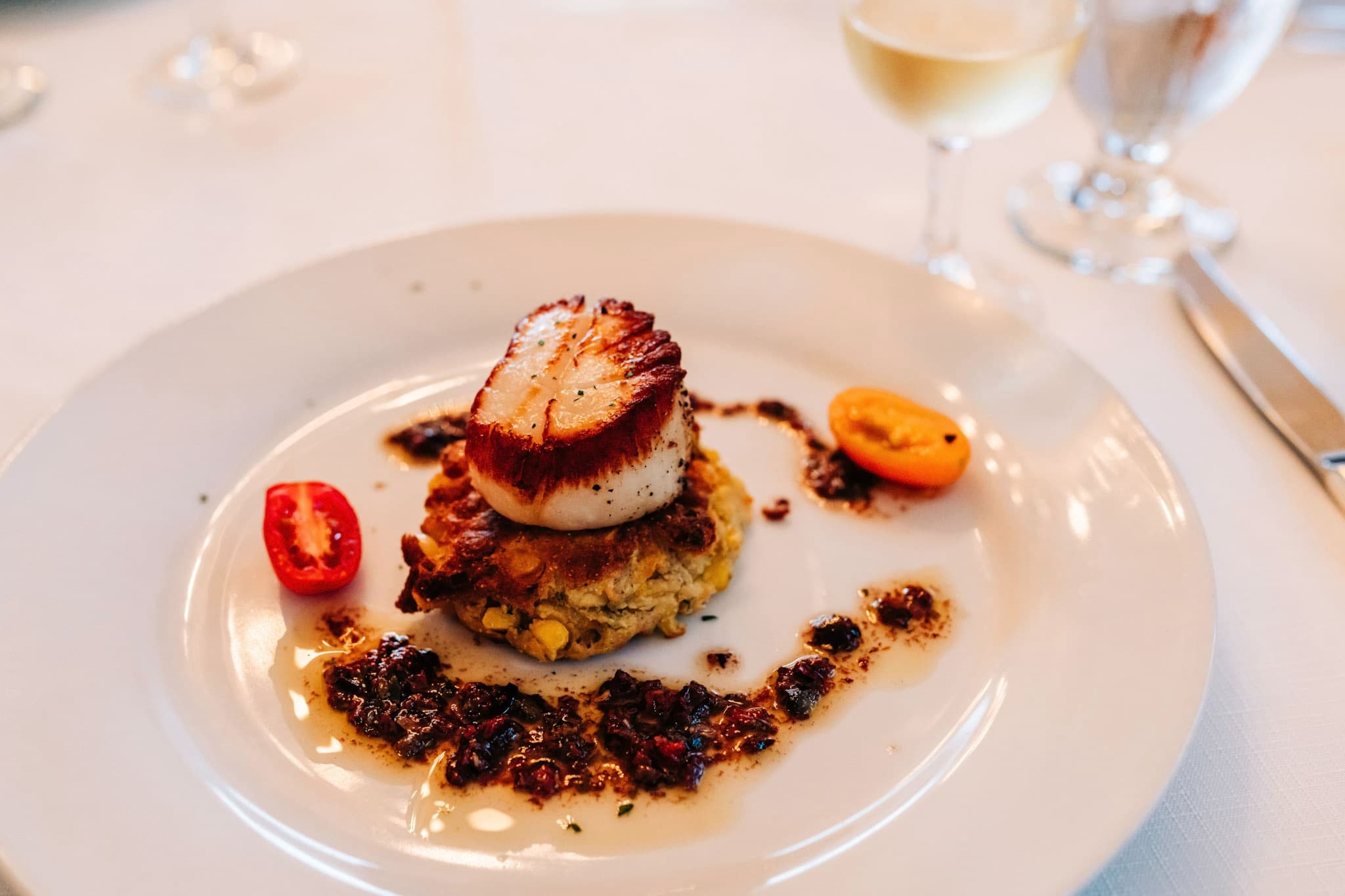 A seared scallop atop a corn cake, garnished with cherry tomatoes and a savory sauce.