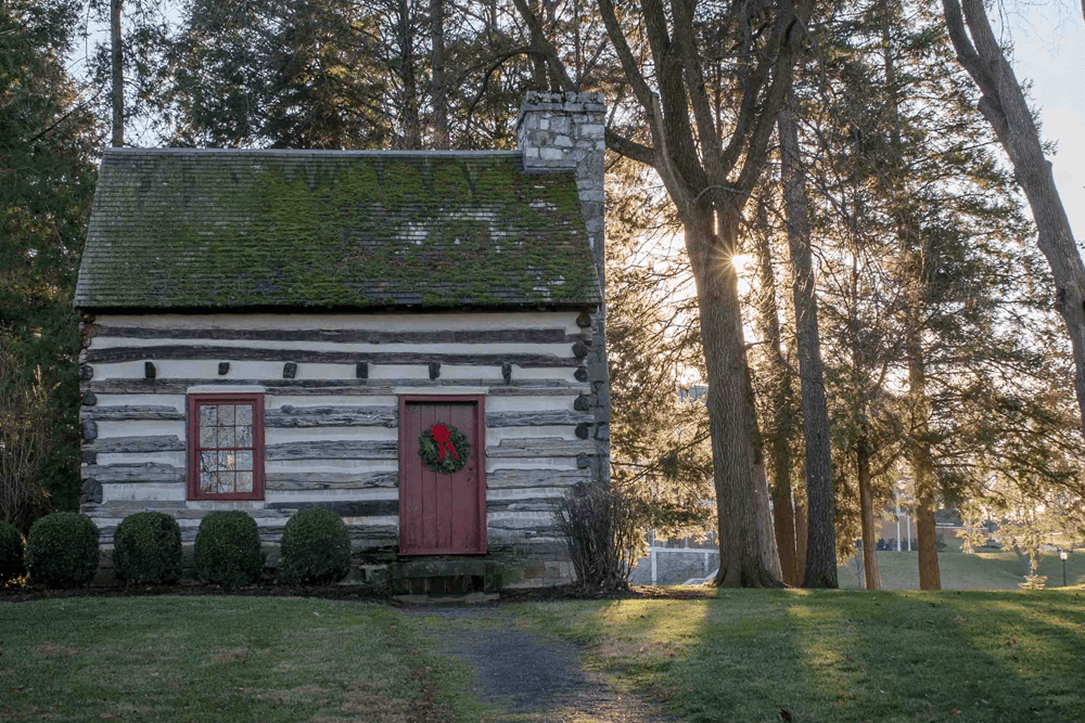 A rustic log cabin adorned with a festive wreath, surrounded by trees and bathed in soft sunlight.