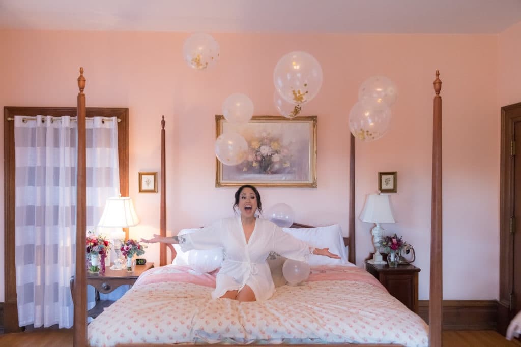A joyful woman in a white robe sits on a bed surrounded by floating balloons in a pastel bedroom.