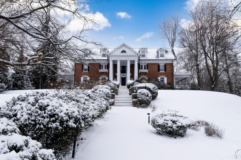 A large brick house with a columned entrance is covered in snow under a bright blue sky.