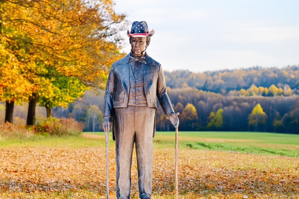 A statue of a man wearing a hat adorned with the American flag stands in an autumn landscape.
