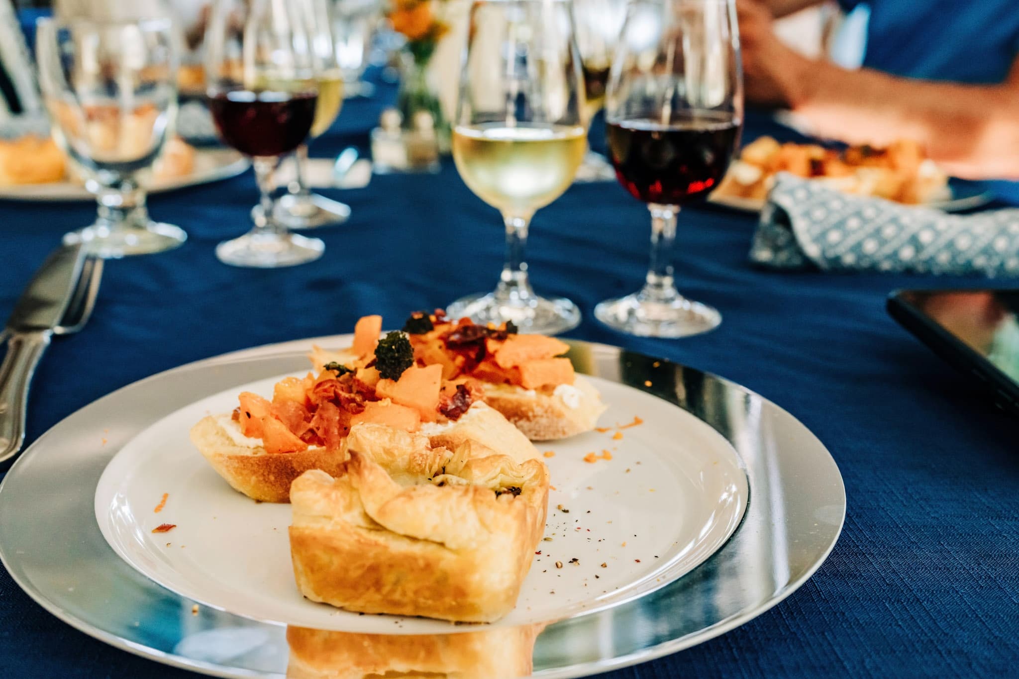 A plated appetizer featuring flaky pastry topped with diced ingredients, surrounded by various glasses of wine on a blue tablecloth.
