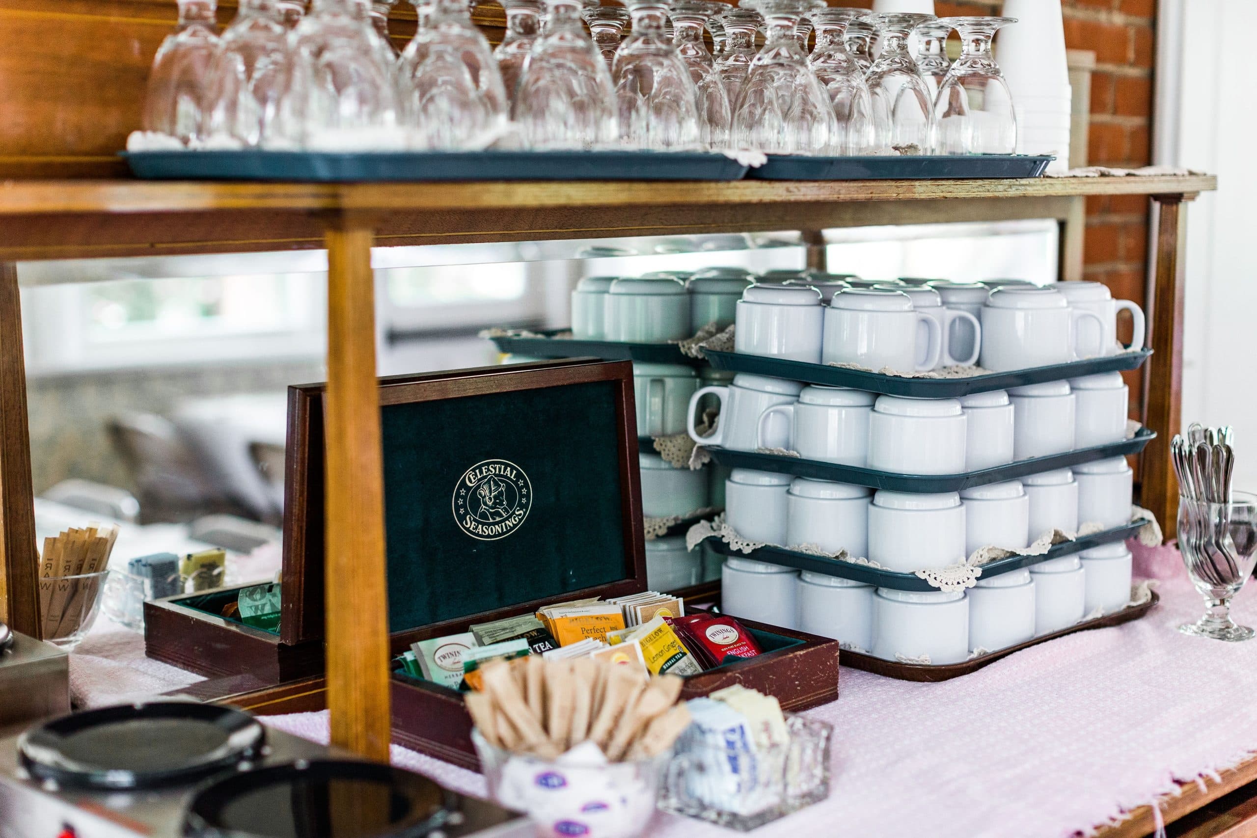 A wooden shelf displaying glassware, stacked mugs, and a box of assorted tea and coffee supplies.
