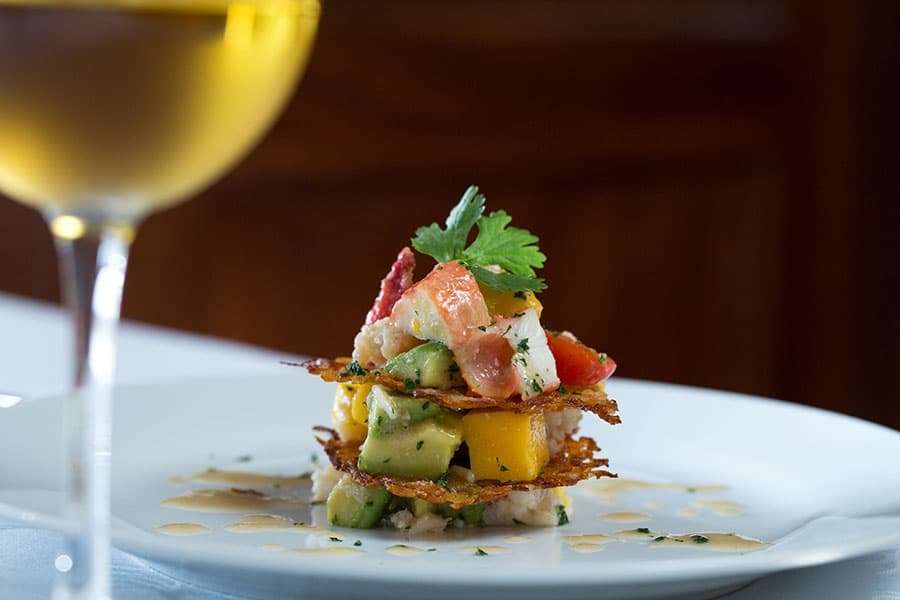 A beautifully arranged dish of layered mango, avocado, and shrimp garnished with cilantro, served beside a glass of white wine.