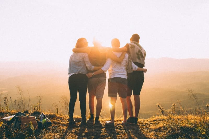 A group of four friends stands together, embracing each other against a sunset backdrop.