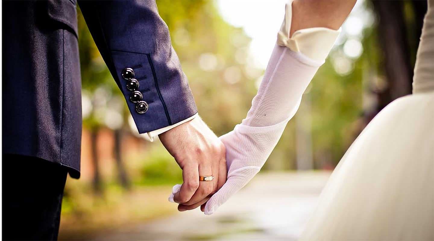 A groom and bride hold hands, showcasing their wedding rings in a serene outdoor setting.