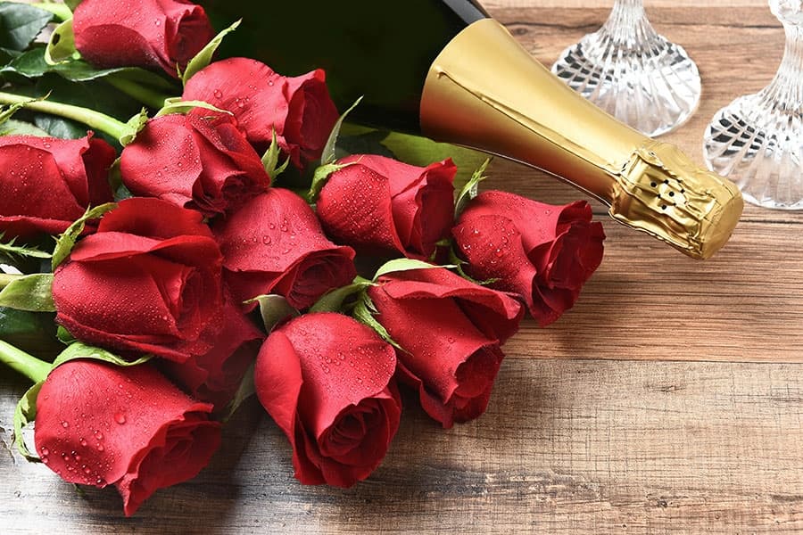 A bouquet of red roses beside a bottle of champagne and two crystal glasses on a wooden surface.