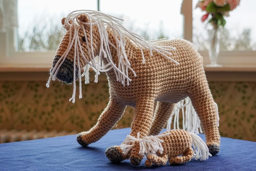 A crocheted horse and a foal are displayed on a blue tablecloth near a window.