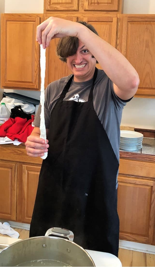 A smiling person in an apron holds a long piece of stretched dough over a pot in a kitchen.