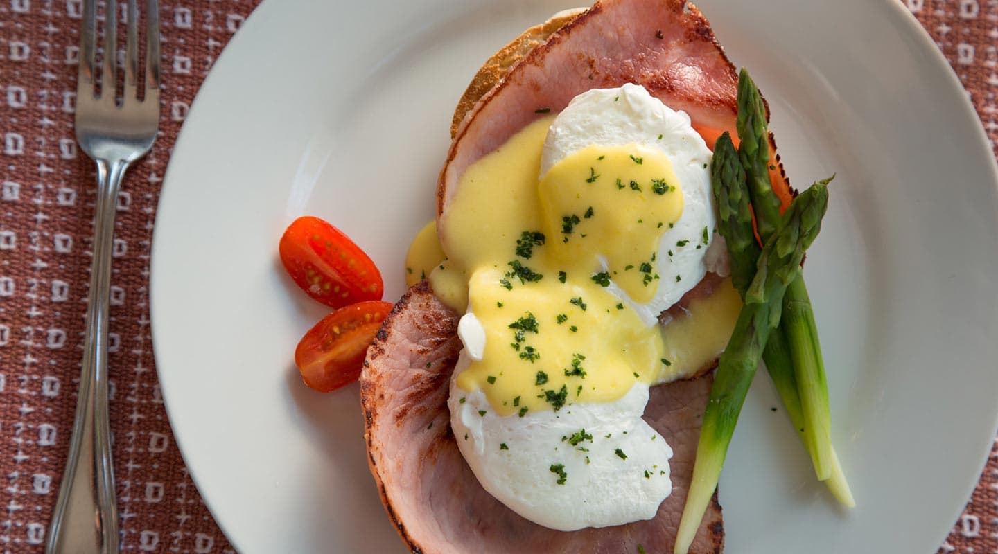 A plate of eggs Benedict with ham, poached eggs, hollandaise sauce, asparagus, and cherry tomatoes.