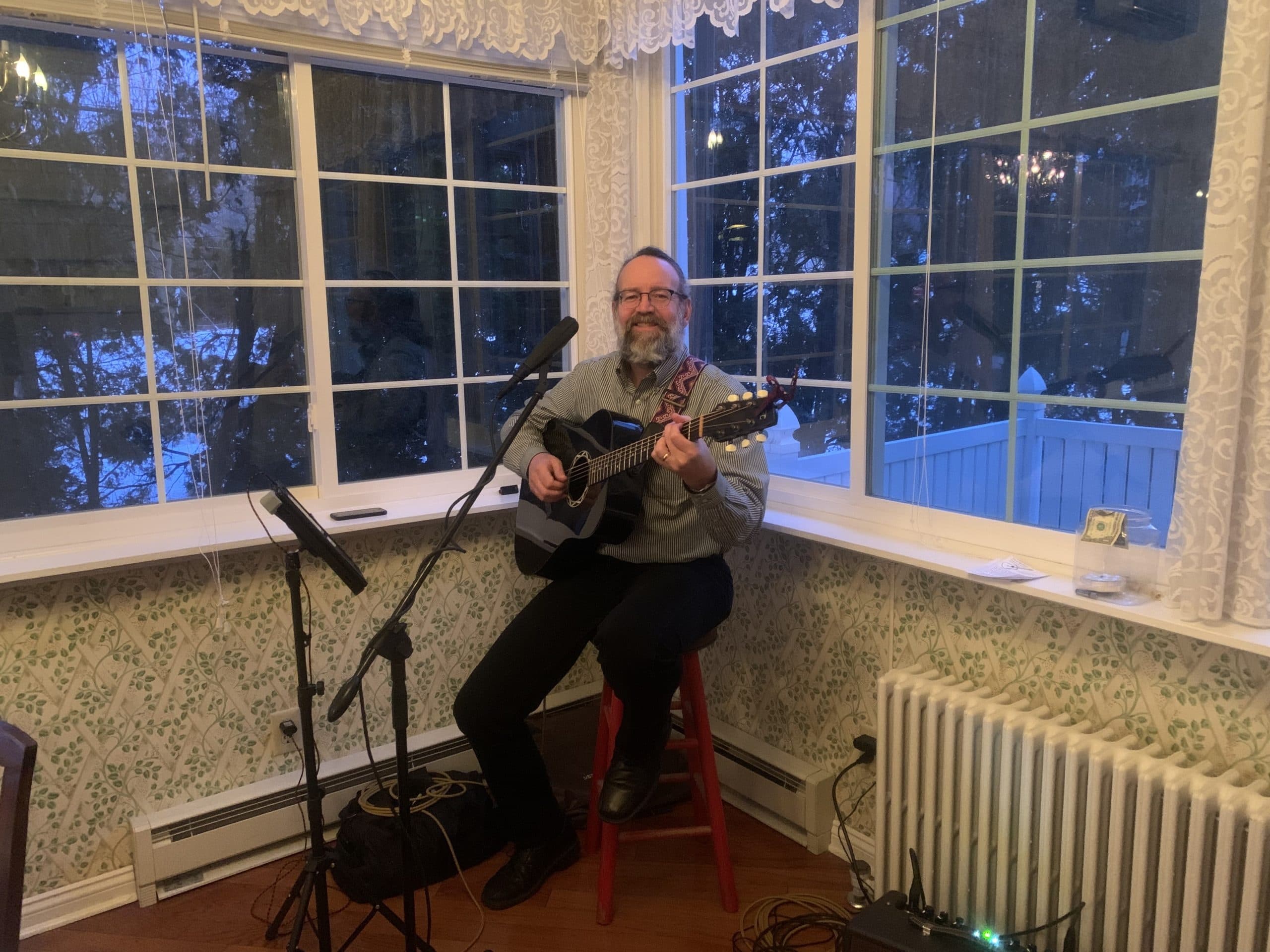 A man plays guitar while seated by a window in a cozy room.