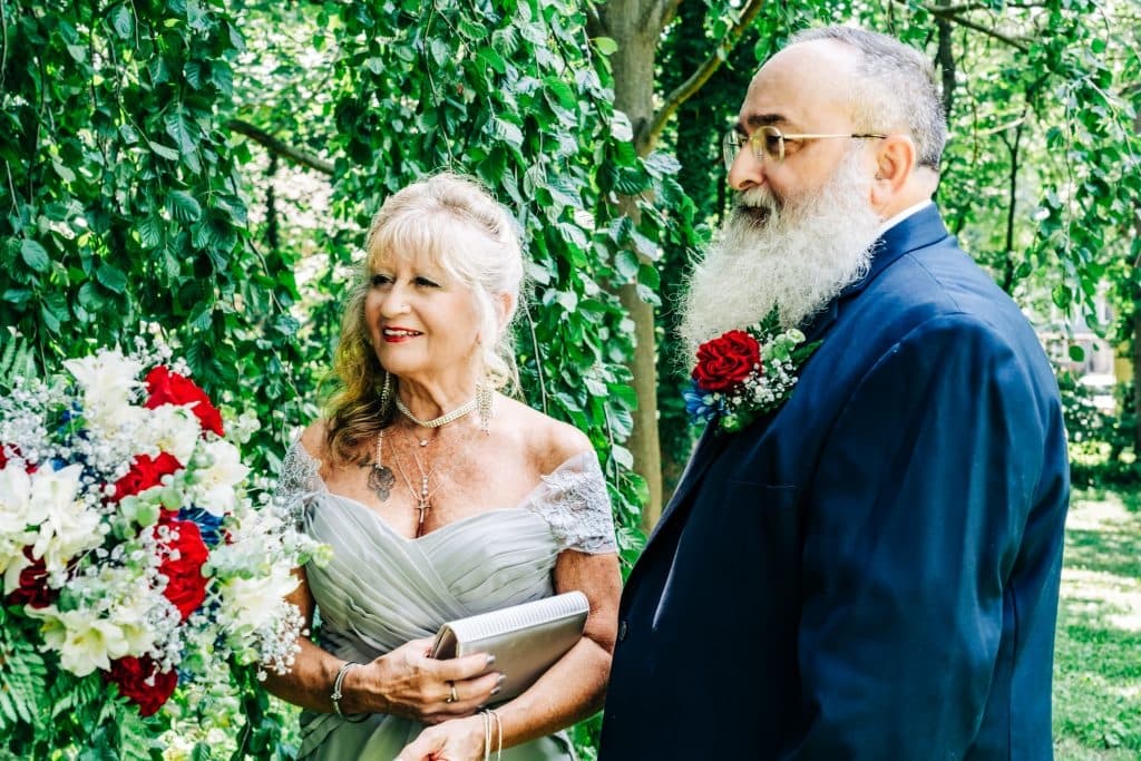 A smiling woman in a gray dress holds a bouquet, standing beside a bearded man in a suit, both in a lush green setting.