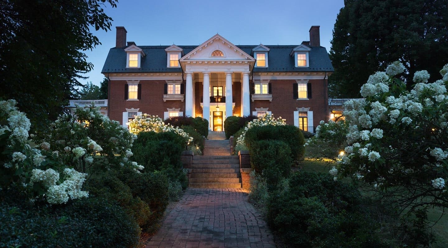 A grand brick mansion illuminated at dusk, surrounded by blooming flowers and lush greenery.