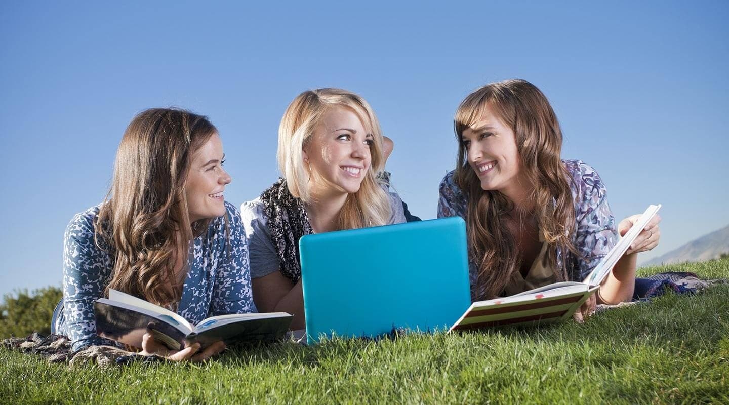 Three young women smiling and studying together on a grassy area, with books and a laptop in front of them.