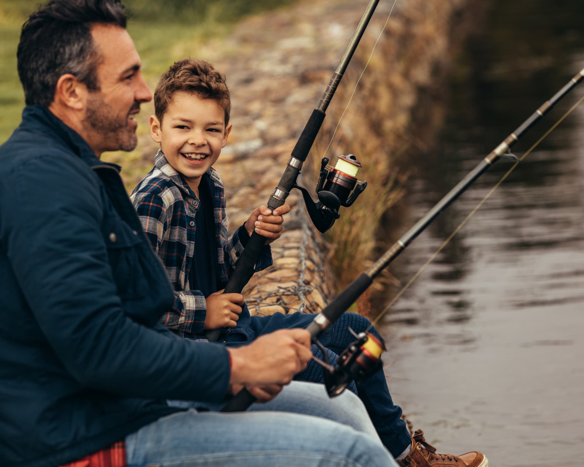 A man and a boy smile while fishing together by a tranquil riverbank.