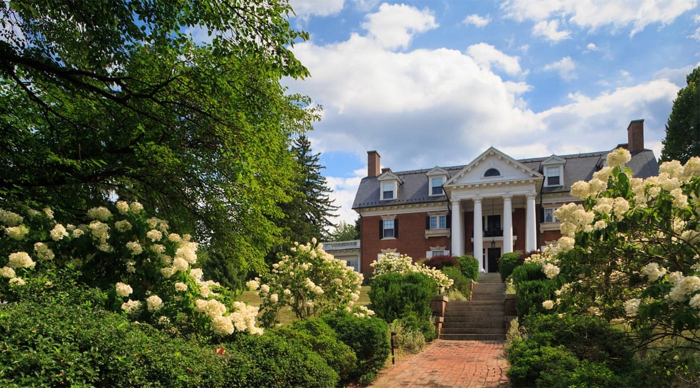 A grand brick mansion surrounded by lush greenery and flowering bushes under a partly cloudy sky.