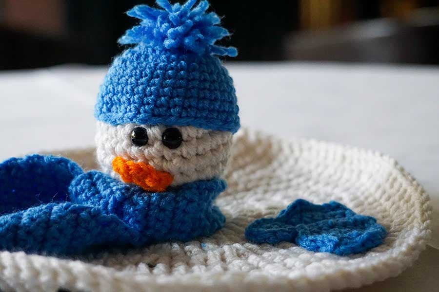 A crocheted snowman wearing a blue hat and scarf sits on a white crocheted plate.