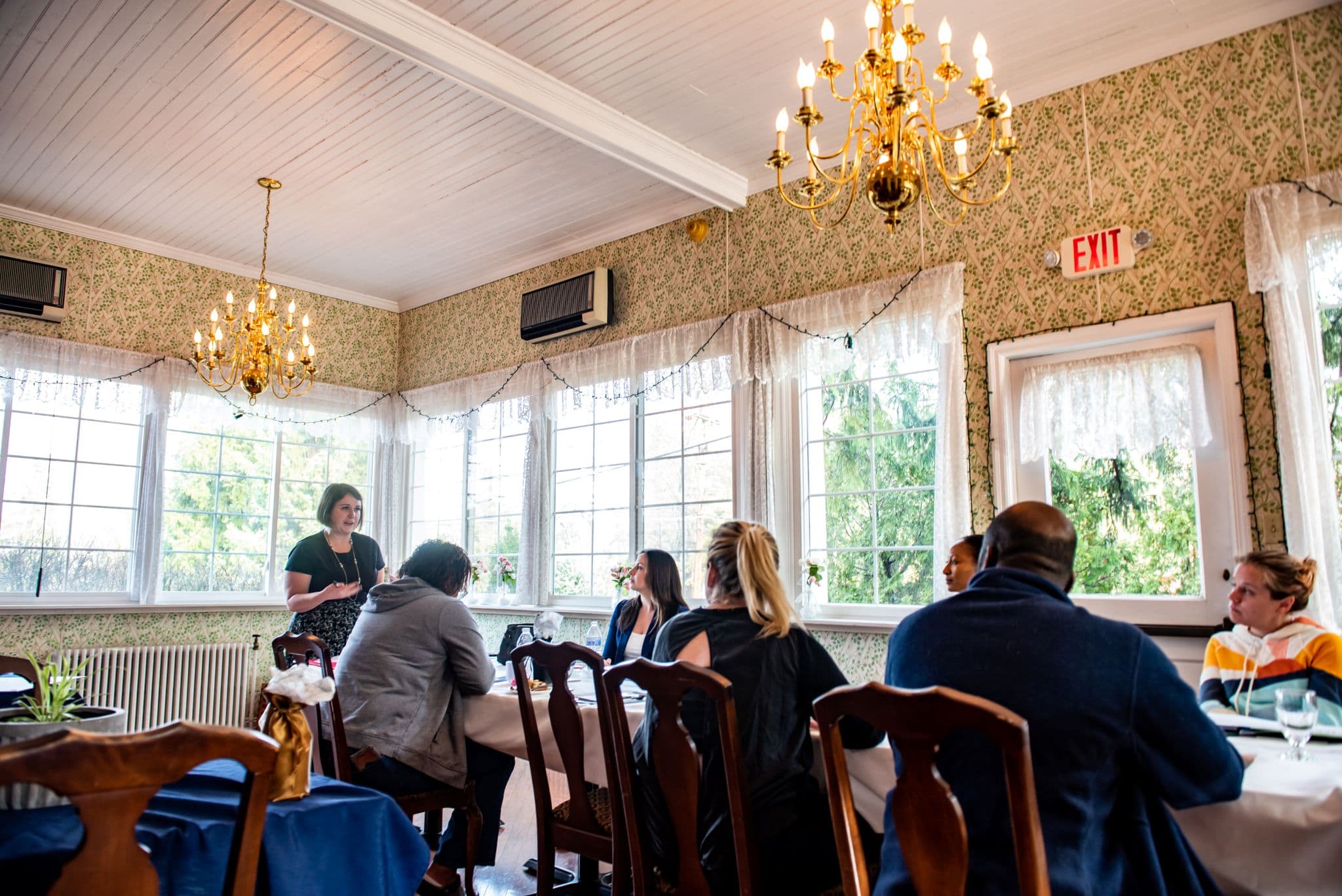 A woman stands and speaks to a group seated at tables in a well-lit restaurant with chandeliers and large windows.