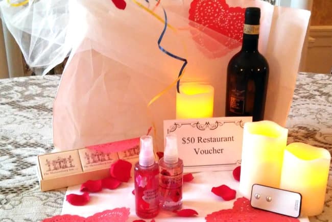 A romantic gift basket featuring a bottle of wine, candles, a $50 restaurant voucher, and scented sprays, surrounded by rose petals.