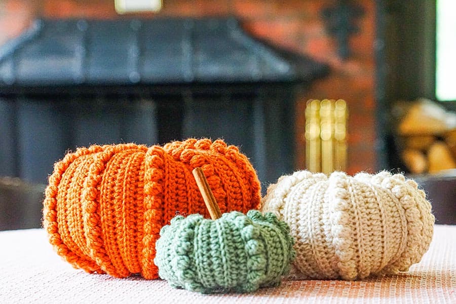 Three crocheted pumpkins in orange, green, and cream sit on a table, with a brick wall and a fireplace in the background.