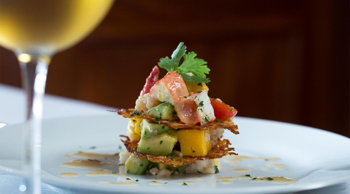 A beautifully plated seafood stack with mango, avocado, and herbs, accompanied by a glass of white wine.
