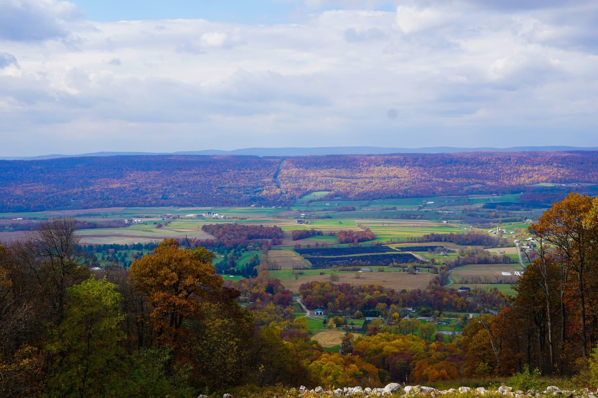 A panoramic view of a colorful autumn landscape, featuring rolling hills and farmland under a cloudy sky.