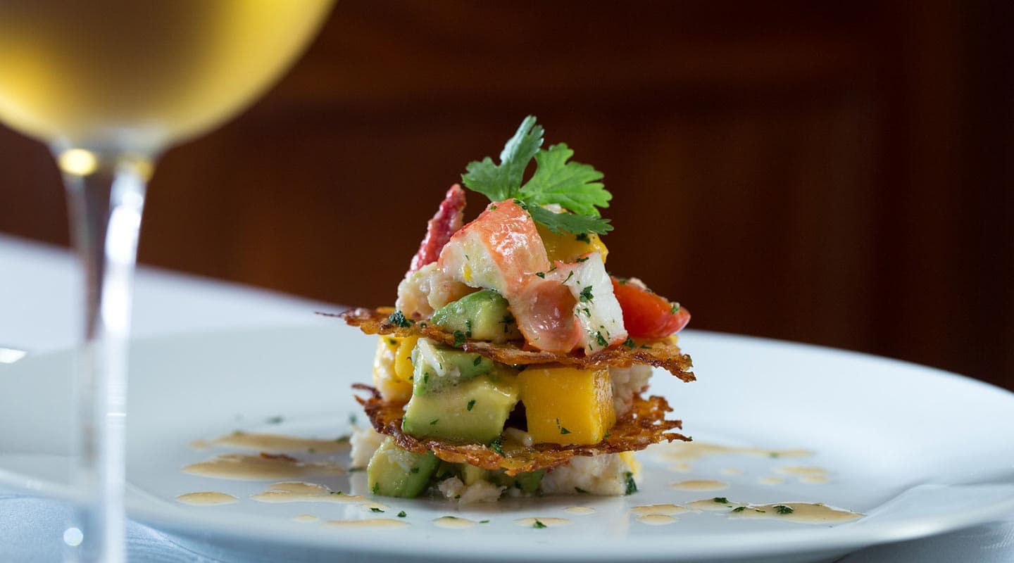 A colorful seafood and avocado tower garnished with herbs, served on a white plate alongside a glass of white wine.