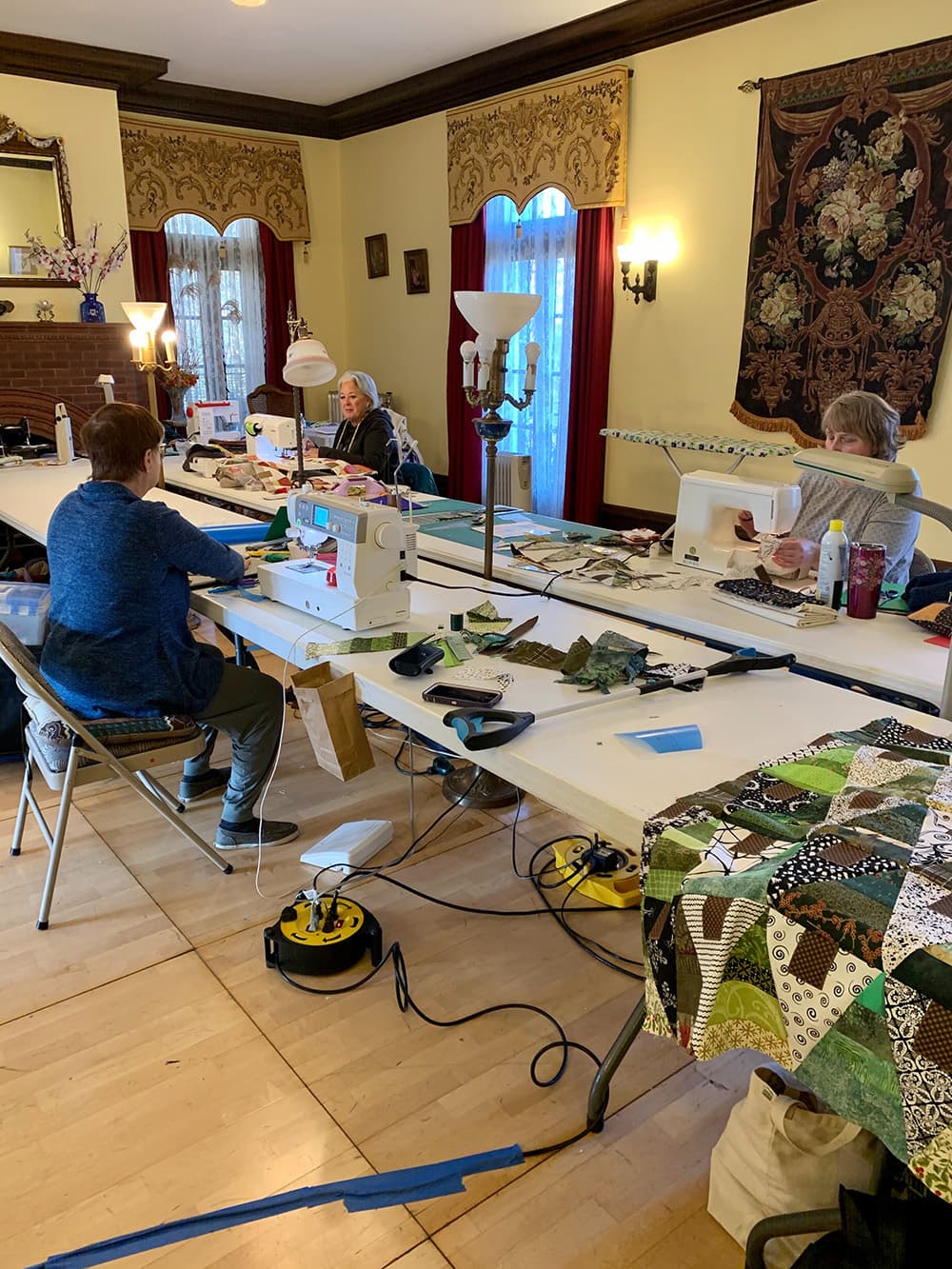 Three people are seated at a long table sewing and quilting in a well-lit room with windows and decorative curtains.