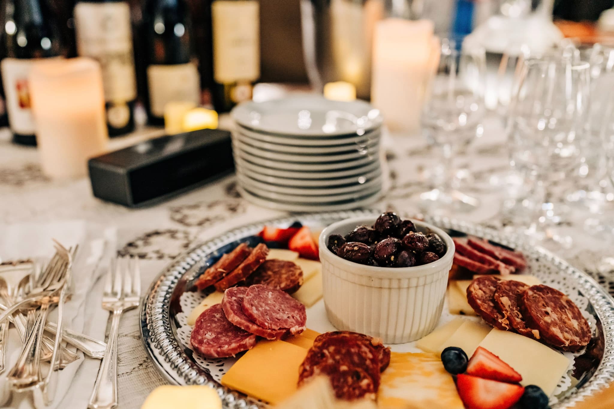 A beautifully arranged platter featuring various cheeses, cured meats, olives, and fresh fruit, set against a backdrop of candles and wine bottles.