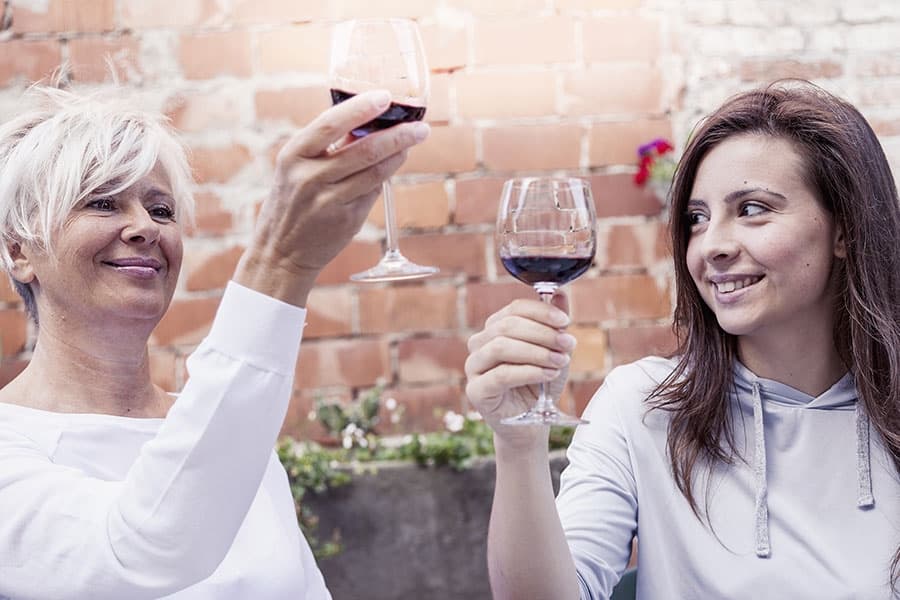 Two women toast with glasses of red wine against a rustic brick background.