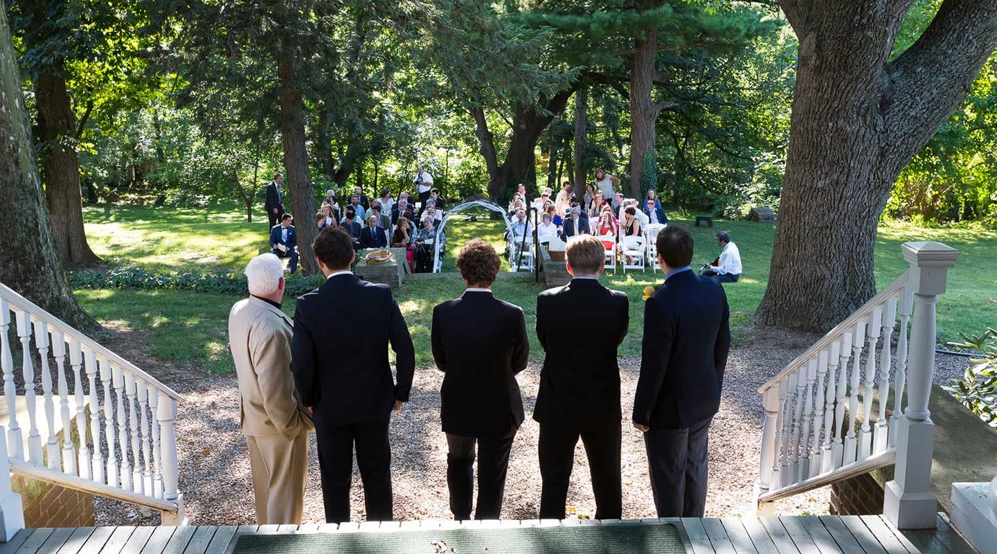 A group of men in suits stands on a staircase overlooking a wedding ceremony surrounded by guests in a lush outdoor setting.
