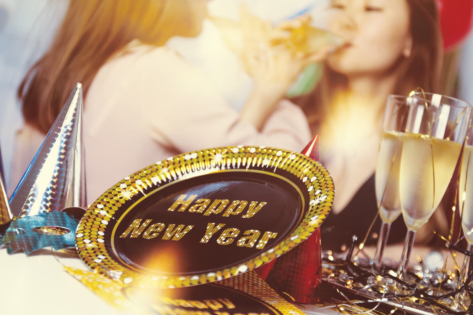 Two women celebrate New Year's Eve with champagne, wearing party hats, surrounded by festive decorations.