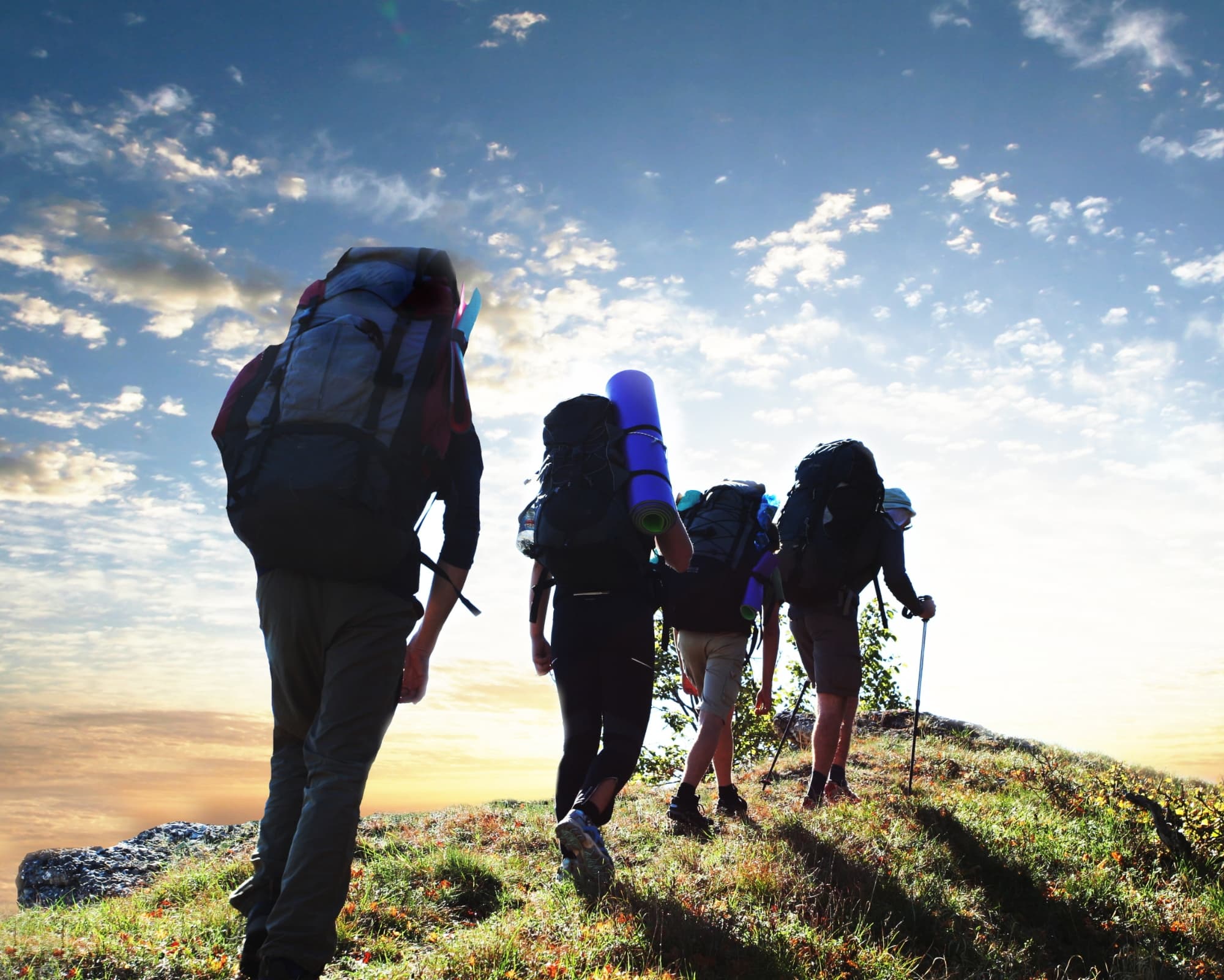 A group of hikers carrying backpacks and sleeping mats ascend a grassy hill against a sky filled with clouds at sunset.