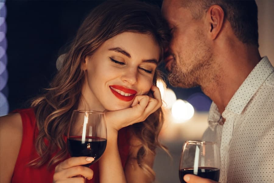 A couple shares a romantic moment, smiling and holding glasses of red wine in a softly lit setting.