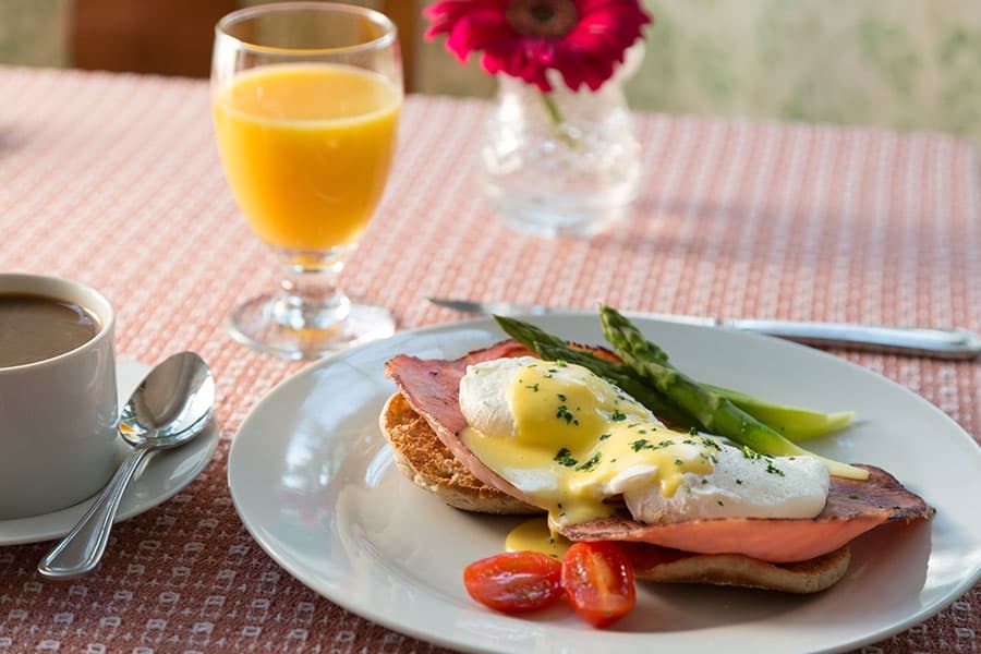 A plate of eggs Benedict with ham, asparagus, and tomatoes, accompanied by a cup of coffee and a glass of orange juice on a patterned tablecloth.
